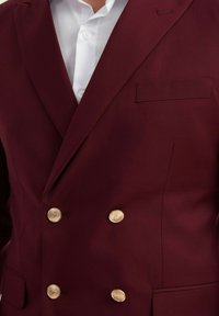 Blazer double boutonné bordeaux en tissu lisse, avec des boutons dorés, une poche poitrine simple et des revers élégants.