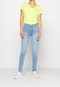 Una maglietta gialla brillante a scollo a V abbinata a jeans skinny leggeri blu chiaro con vita alta, caratterizzati da un frontale con bottoni e sneakers bianche con platfond.