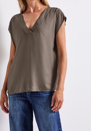 Femme portant un chemisier sans manches vert olive foncé à col en V associé à un jean bleu en denim sur un fond uni.