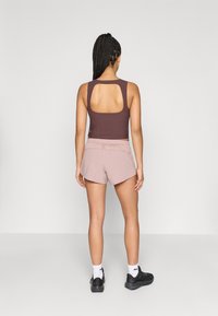 Αθλητική μπλούζα χωρίς μανίκια σε σκούρο καφέ με άνοιγμα στην πλάτη, συνδυασμένη με ροζ σορτς. Το μοντέλο φοράει μαύρα αθλητικά παπούτσια και λευκές κάλτσες αστραγάλου.