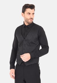 Gilet nero con un intricato motivo testurizzato, caratterizzato da un neckline a V, sei bottoni e una vestibilità su misura. Indossato sopra una camicia nera slim.