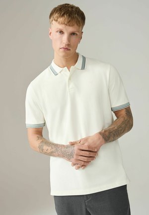 Noor mees lühikeste juustega ja tätoveeringutega mõlemal käsivarrel, kandes valget poloshirti triibulise krae ja varrukalõigetega.