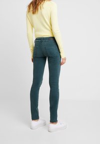 Mujer vestida con jeans ajustados color verde azulado, blusa de manga larga amarilla y zapatillas blancas, de pie contra un fondo claro y liso.