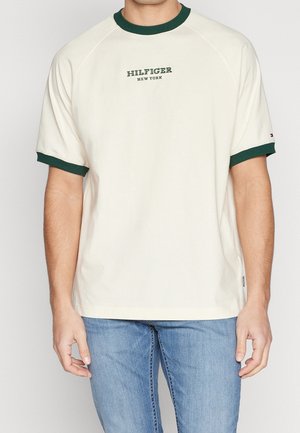 Mand iført en cremefarvet T-shirt med korte ærmer med grøn krave og ærmekanter, tekst "HILFIGER NEW YORK" på brystet og blå jeans.
