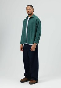 Grünes Hemd über einem weißen T-Shirt, kombiniert mit dunkelblauen Weitbein-Jeans und braunen Schnürstiefeln.