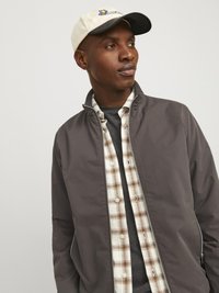 Gray zip-up jacket with a high collar, layered over a checkered beige and brown shirt, worn with a light-colored cap featuring a logo.