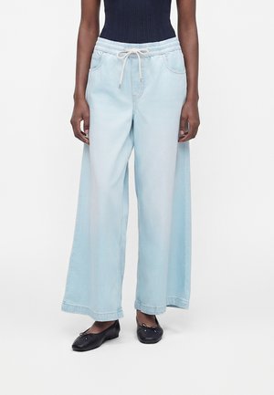 Osoba nosiaca svetlomodré široké rifle so šnúrkou v páse a čierne balerínky, stojaca na bielom pozadí.