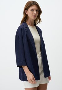 Donna che indossa un blazer aperto blu navy, una maglietta beige e pantaloni corti bianchi a vita alta, in piedi davanti a uno sfondo chiaro e semplice.