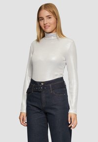 Silberner, langärmeliger Rollkragenpullover mit glattem, glänzendem Material, kombiniert mit dunkelblauen Mom-Jeans mit hoher Taille, die eine sichtbare Gürtelschlaufe und Knopfdetails aufweisen.