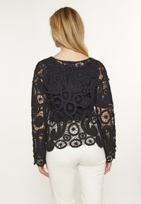 Cardigan din croșetă negru, cu modele intricate, mâneci lungi și un design cu spatele deschis, asortat cu o piesă de îmbrăcăminte de jos de culoare deschisă.
