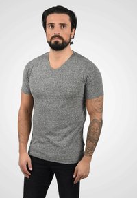 Camiseta gris de manga corta y cuello en V, hecha de una tela texturizada. Llevada sobre unos jeans negros, con un tatuaje visible en el brazo izquierdo.