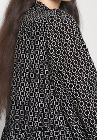 Schwarzes Kleid mit langen Ärmeln, das ein geometrisches weißes Kästchenmuster aufweist, einen hohen Kragen und einen Rüschensaum hat. Weicher Stoff.