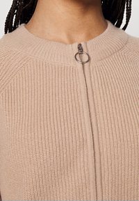 Beige knit sweater with a round neck. Features a front zipper and ribbed texture along the body and sleeves. Silver hardware detail at the zip.