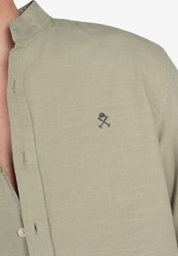 Camisa de color verde claro con cuello, fabricada en un tejido texturizado, que presenta un emblema bordado en color marino. Detalles de botones visibles en la tapeta delantera.