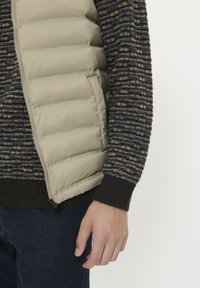 Beige pufferväst med ett kviltat mönster i lager över en flerfärgad stickad tröja med mörka och ljusa ränder. Mörka jeans syns nedanför.