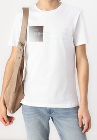 Osoba oblečená v bílé grafické tričko, světle modré džíny a nese béžovou semišovou tašku přes rameno na levé paži.