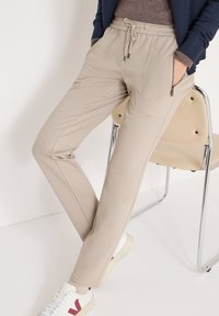 Beige stretch joggers met een taillekoord, zijzakken en een gladde textuur. Gecombineerd met witte sneakers en een donker vest.