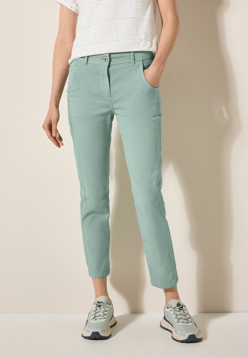 Personne portant un pantalon slim vert clair, un haut blanc à manches courtes perforé et des baskets vert clair, debout devant un fond beige.