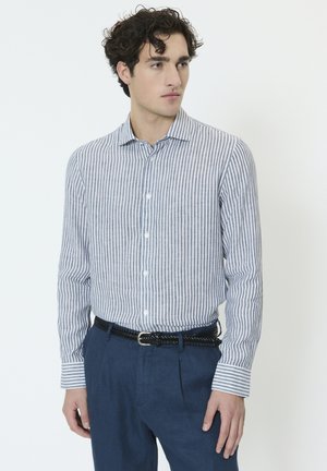 Nooruk lokkis juustega mees, kes kannab sinist ja valget vertikaaltriipudega särki, mis on pistetud sinistesse pükstesse, millel on must punutud vöö, vaadates küljele.