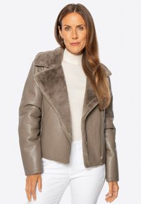 Grijze leren jas met een zacht nepbont kraag, ritssluiting en lange mouwen, gedragen over een lichte trui en witte broek.