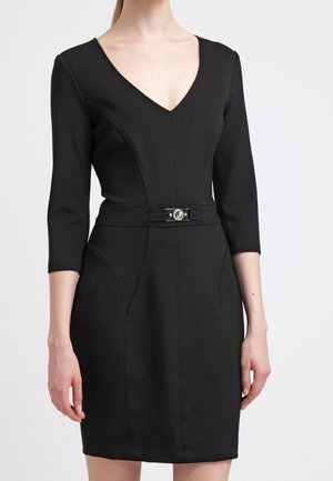 Robe noire près du corps avec manches trois-quarts, encolure en V et ceinture à la taille avec boucle métallique, portée par une personne sur un fond uni.