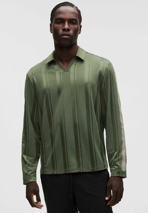 Uomo con capelli intrecciati indossa una camicia verde a maniche lunghe con righe verticali e colletto, abbinata a pantaloni neri, in piedi davanti a uno sfondo uniforme.