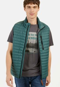 Teal puffervest med horisontale quiltede linjer, en høj krave og en skrå lynlåslomme, båret over en grå grafisk t-shirt.