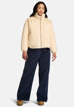 Lichtbeige pufferjack met een hoge kraag, ritssluiting en gestikte panelen, gecombineerd met donkerblauwe wijde broekspijpen en tan laarzen.