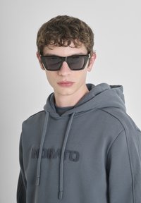 Felpa grigia con cappuccio e cordini regolabili, logo ricamato sul petto, abbinata a occhiali da sole neri a forma quadrata. Superficie liscia.