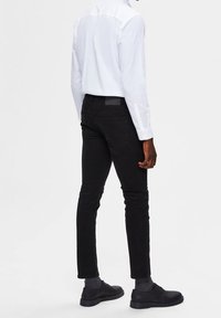 Svart slim-fit byxor med en slät textur, med bakfickor och en diskret märkesetikett. Kombineras med en vit skjorta med knappar.