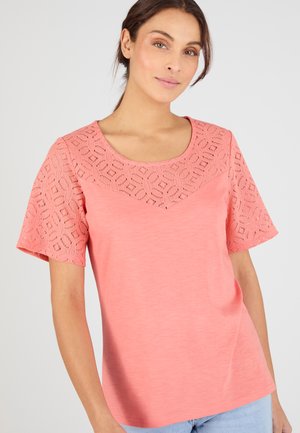 Mujer con una blusa rosa coral de manga corta con detalles geométricos de encaje en las mangas y el pecho superior, combinada con jeans azul claro.