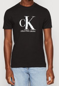 Černé bavlněné tričko s bílým logem "CK" a textem "Calvin Klein Jeans" pod ním. Krátké rukávy, kulatý výstřih a volný střih.