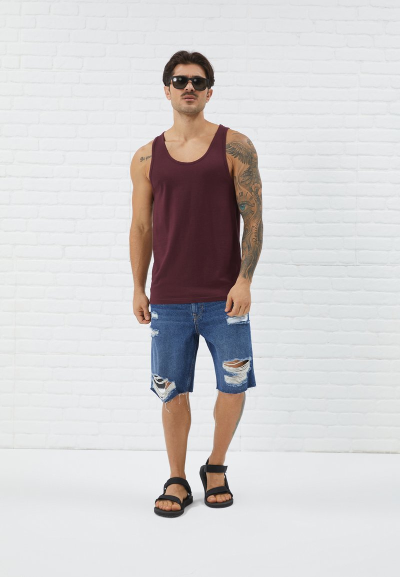Maroon tank top, sortit tummat denim-housut, joissa on risat reunat, mustat sandaalit ja aurinkolasit. Malli seisoo valkoista tiiliseinää vasten.