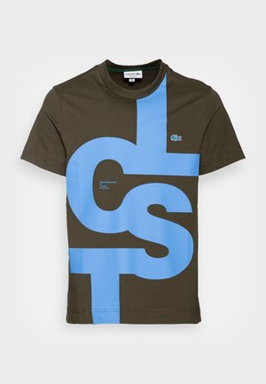 T-shirt en coton vert olive avec de grandes inscriptions bleues, manches courtes et encolure ronde. Comprend un petit logo Lacoste sur la poitrine.