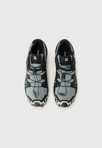Baskets de sport noires et grises avec une surface texturée, un design sans lacets et une semelle blanche contrastée ornée de motifs en zigzag.