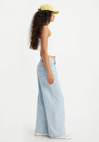 Jeans a gamba larga di un blu chiaro con vita alta, indossati con un top corto bianco e un cappellino giallo, mostrati in profilo su uno sfondo neutro.