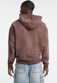Felpa con cappuccio marrone dal taglio rilassato, con polsini e orlo a coste, mostrata dal retro, abbinata a pantaloni cargo azzurri chiari.