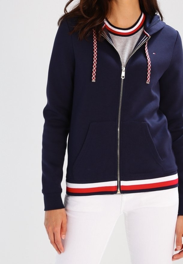 Haut à zip marine avec doublure intérieure grise, accents rayés rouge et blanc sur les poignets et l'ourlet, équipé d'un cordon de serrage et de poches avant.