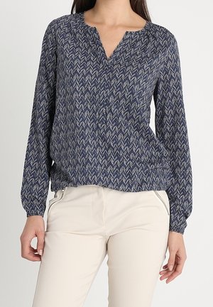 Femme portant un chemisier à manches longues bleu marine à motifs, col en V, et un pantalon crème avec des détails zippés près des hanches.
