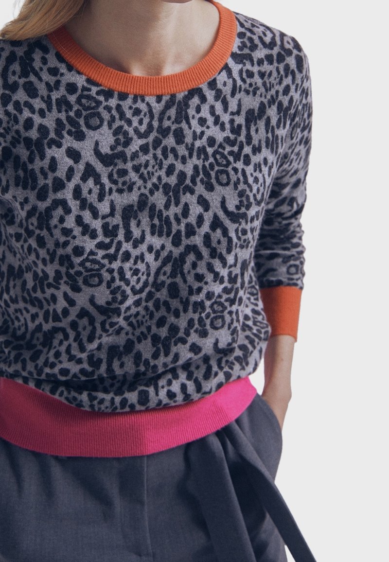 Pull imprimé léopard gris avec des taches noires, col et poignets côtelés orange, et ceinture côtelée rose. Texture de tissu douce.