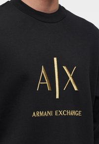 T-shirt noir à col rond avec logo "A|X" brodé en doré et texte "ARMANI EXCHANGE" sur la poitrine.