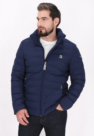 Marineblå pufferjakke med quiltet tekstur og lynlås. Har en høj krave og logo på venstre bryst, parret med mørke denimjeans.