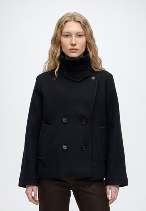 Une femme présente un manteau noir croisé à col montant et un pantalon foncé sur un fond clair et uni.