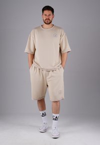 Man standing with hands in pockets wearing beige oversized shorts and T-shirt, white socks with black text, and white sneakers on gray background.