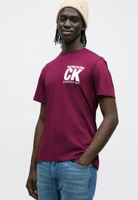 Bordowa bawełniana koszulka z białym logo „CALVIN KLEIN CK Established 1968”. W zestawie z jasnoniebieskimi dżinsami. Krótkie rękawy, okrągły dekolt.