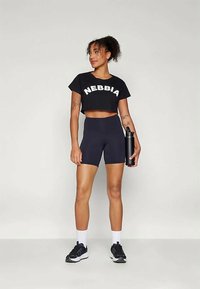 Camiseta preta cropped com o logo "NEBBIA", combinada com calções de cor azul-marinho. O modelo segura uma garrafa de água preta, vestindo meias brancas e sapatilhas pretas.