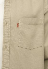 Μπεζ πουκάμισο με κουμπιά και τσέπη στο στήθος με μικρή κόκκινη ετικέτα Levi's, φορεμένο πάνω από λευκό ριμπ ύφασμα.