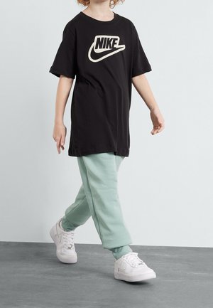 Enfant marchant portant un t-shirt noir Nike, un pantalon de survêtement vert clair et des baskets blanches Nike sur un sol gris devant un fond blanc.