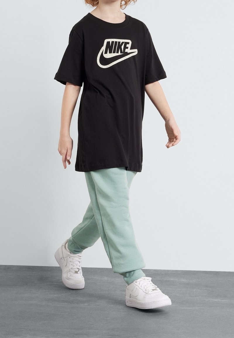 Barn som går i en svart Nike-t-shirt, ljusgröna träningsbyxor och vita Nike-sneakers på ett grått golv mot en vit bakgrund.