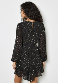 Schwarzes Chiffonkleid mit langen Ärmeln, Rüschenausschnitt und goldenen Sternenmustern. Mit einem Schlüsselaugen-Design im Rücken und einem elastischen Bund.